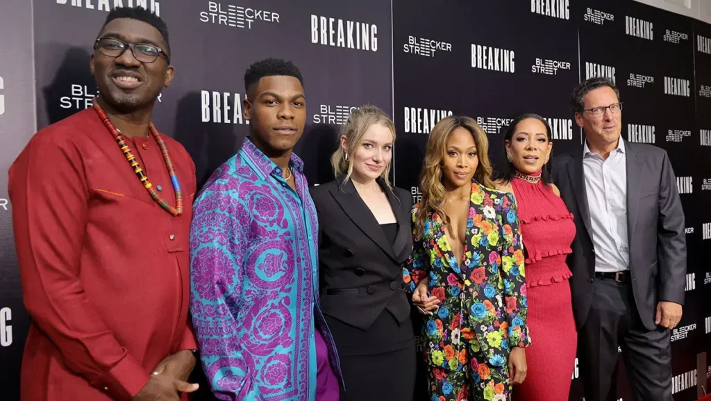 Skupinová fotografie na premiéře filmu "Breaking" od Bleecker Street. Zleva doprava: Muž v červeném tradičním africkém oděvu s korálovým náhrdelníkem, herec v modrofialové košili s výrazným vzorem, herečka v černém saku, herečka v barevném květinovém kostýmu, herečka v červených šatech a muž v šedém obleku s bílou košilí. Všichni stojí před černou stěnou s logy filmu "Breaking" a společnosti "Bleecker Street