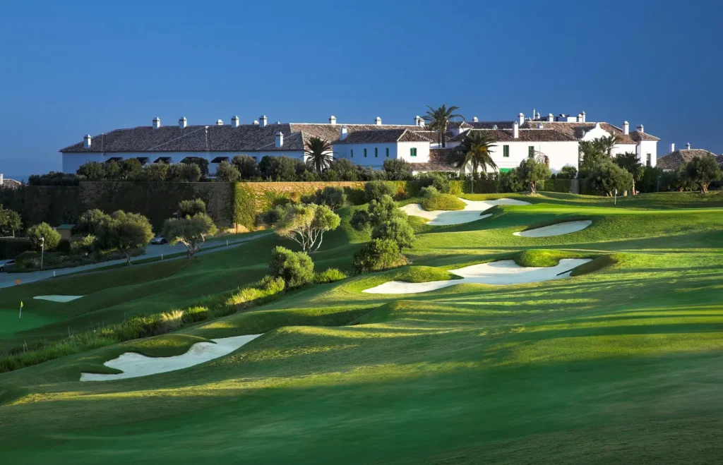 Obrázek zobrazuje golfové hřiště Finca Cortesin ve Španělsku. Hřiště je obklopené krásnou zelení a dokonale udržovanými fairwayi a pískovými bunkry. V pozadí se nachází elegantní bílé budovy s tradičními střechami, které dodávají místu šarm středomořské architektury. Krajina je ozdobena stromy a keři, které harmonicky ladí s okolním prostředím. Osvětlení vytváří jemné stíny na trávníku, což dodává celé scenérii klidnou a idylickou atmosféru, ideální pro golfovou hru. Modrá obloha bez mráčku dokonale doplňuje tento malebný výhled.