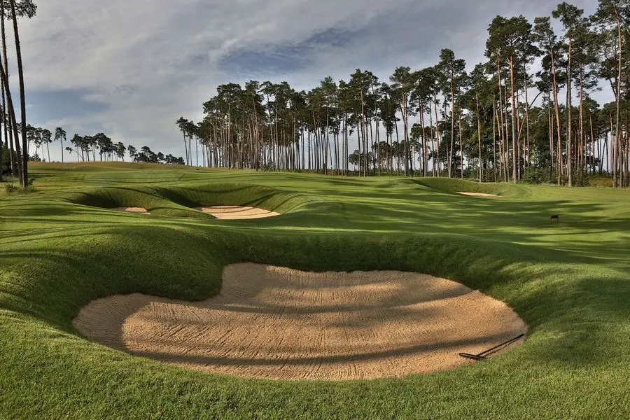 Golfové hřiště obklopené vysokými stromy, s dobře udržovanými fairwayemi a pískovými bunkery v popředí, pod jasnou oblohou.