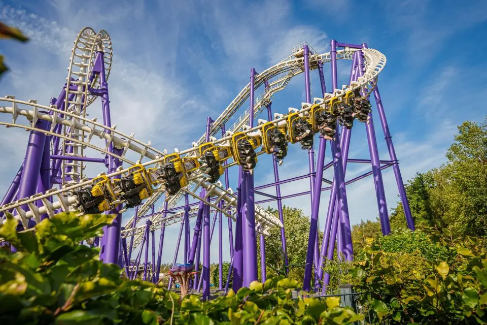 Fotografie zachycuje adrenalinovou horskou dráhu v zábavním parku Heide Park v Německu. Horská dráha má výraznou fialovou konstrukci s bílými kolejemi a žlutými vagónky. Na dráze vidíme návštěvníky, kteří si užívají rychlou a vzrušující jízdu plnou smyček a zatáček. V popředí jsou vidět zelené keře a stromy, které přidávají na přírodní atmosféře parku. Nebe je modré s několika bílými mraky, což naznačuje pěkné a slunečné počasí.