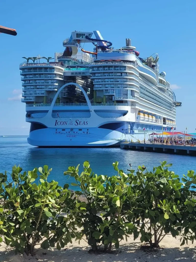 Ikonická výletní loď Icon of the Seas od Royal Caribbean zakotvená v přístavu, s jasně modrou oblohou a pohledem na její velkolepé paluby a balkony, se zelení v popředí.