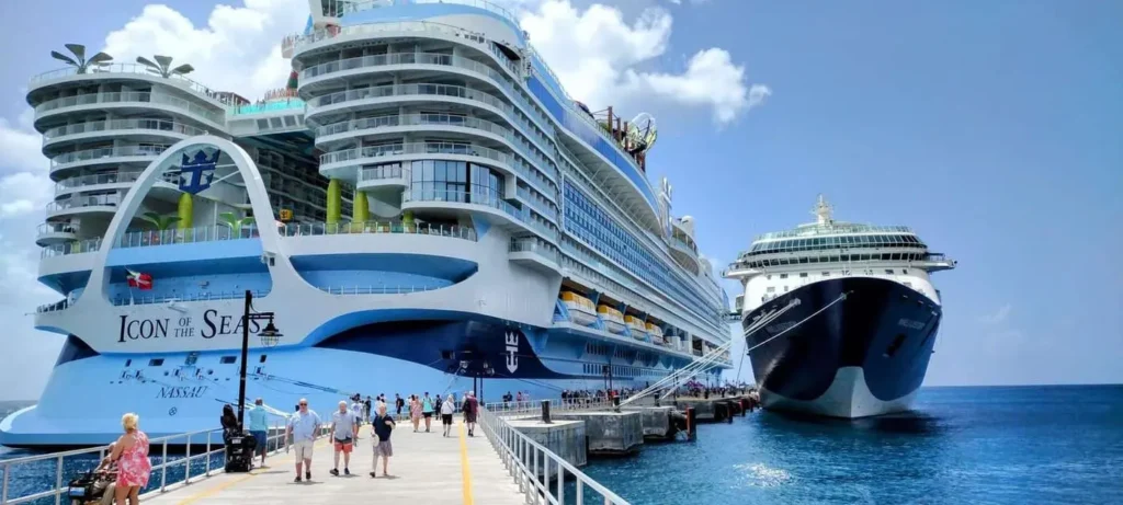 Fotografie výletní lodi Icon of the Seas od Royal Caribbean, zakotvené vedle další výletní lodi, s turisty procházejícími po molu pod modrou oblohou.