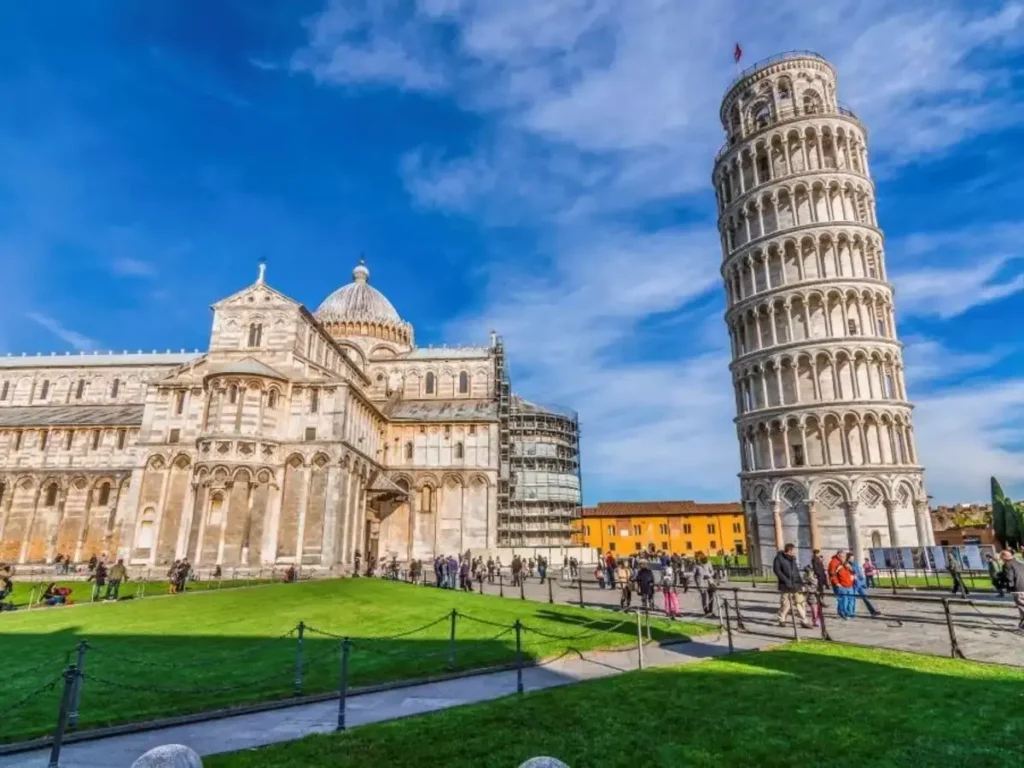 Fotografie zachycuje slavnou Šikmou věž v Pise, Itálie, spolu s katedrálou Panny Marie Nanebevzaté (Duomo di Pisa). V popředí je zelený trávník, na kterém se pohybují turisté, kteří si užívají pohled na tuto ikonickou stavbu. Věž, známá svým výrazným náklonem, se tyčí vedle majestátní katedrály, která je zdobena bohatými architektonickými detaily a kupolí. Na pozadí je modrá obloha s několika mraky, což dodává scéně jasnost a živost. Celkově fotografie představuje jedno z nejznámějších turistických míst v Itálii, které láká návštěvníky z celého světa.