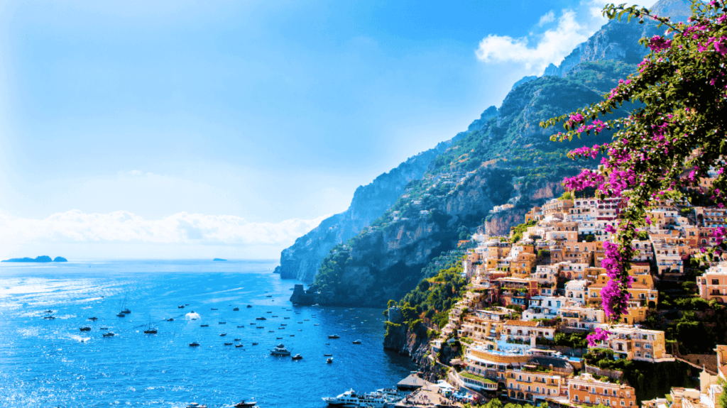 Fotografie zachycuje nádherný výhled na pobřežní město Positano v Itálii. Město je postavené na strmých svazích a je charakteristické svými barevnými domy, které se kaskádovitě táhnou dolů k tyrkysově modrému moři. V popředí jsou vidět fialové květy, které přidávají na živosti a barevnosti scény. V moři je mnoho malých lodí a člunů, které dotvářejí atmosféru přímořského letoviska. Na pozadí se tyčí zelené kopce a hory, které poskytují dramatický kontrast k modré obloze a moři. Celková atmosféra je klidná a malebná, ideální pro dovolenou plnou relaxace a krásných výhledů.