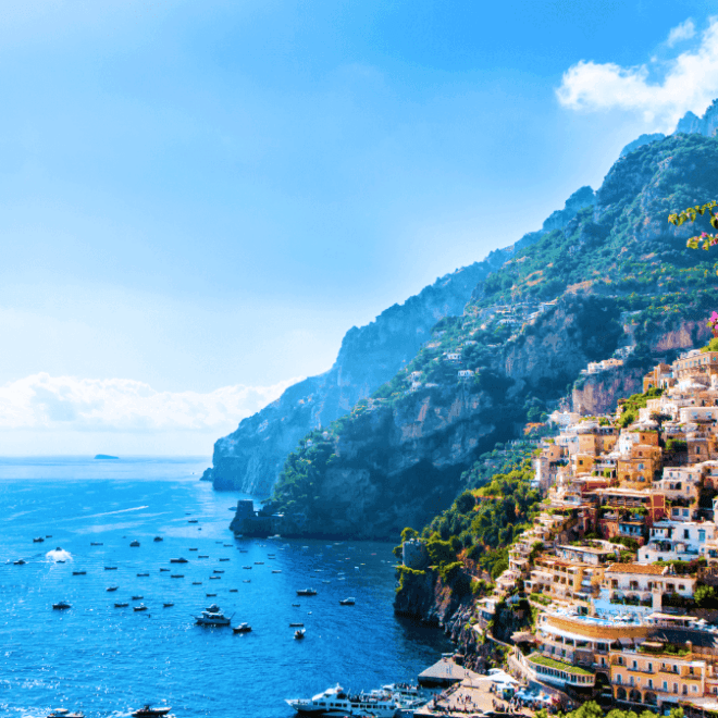 Fotografie zachycuje nádherný výhled na pobřežní město Positano v Itálii. Město je postavené na strmých svazích a je charakteristické svými barevnými domy, které se kaskádovitě táhnou dolů k tyrkysově modrému moři. V popředí jsou vidět fialové květy, které přidávají na živosti a barevnosti scény. V moři je mnoho malých lodí a člunů, které dotvářejí atmosféru přímořského letoviska. Na pozadí se tyčí zelené kopce a hory, které poskytují dramatický kontrast k modré obloze a moři. Celková atmosféra je klidná a malebná, ideální pro dovolenou plnou relaxace a krásných výhledů.