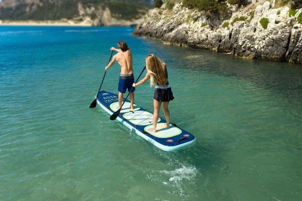 Dva lidé pádlují na rodinném paddleboardu Aqua Marina Super Trip na klidném modrém moři poblíž skalnatého pobřeží.