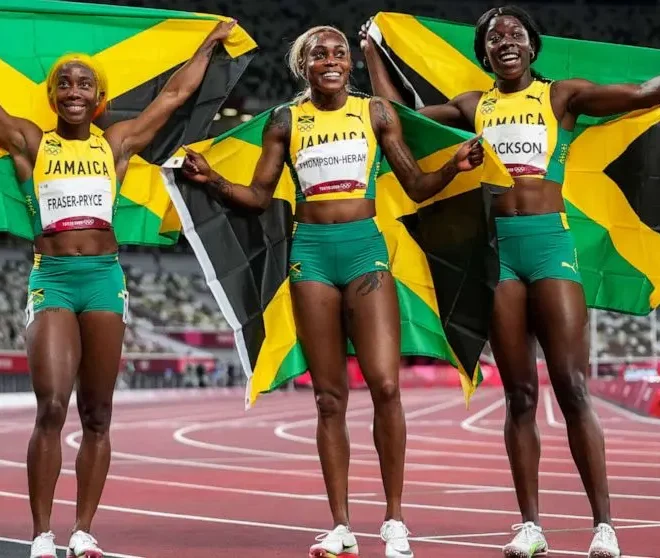 Jamajské sprinterky Shelly-Ann Fraser-Pryce, Elaine Thompsonová-Herahová a Shericka Jackson oslavují společně s jamajskými vlajkami po závodě na atletické dráze.