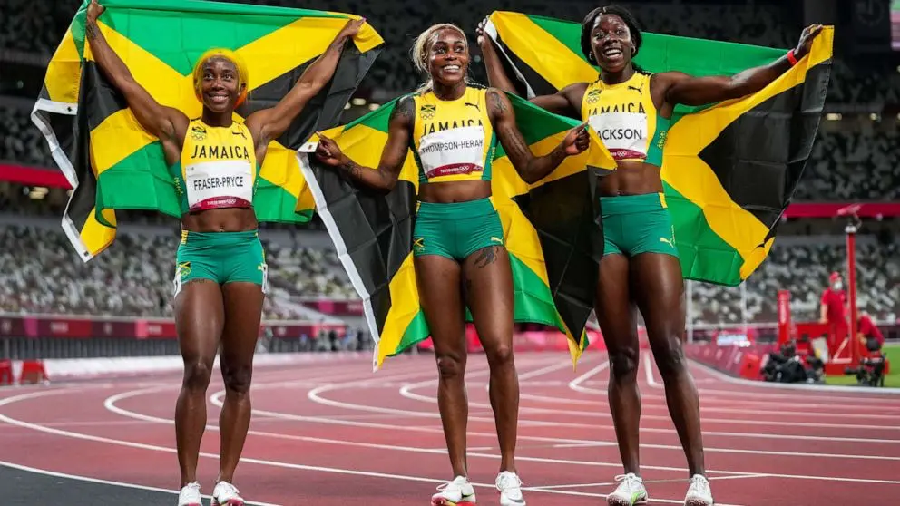 Jamajské sprinterky Shelly-Ann Fraser-Pryce, Elaine Thompsonová-Herahová a Shericka Jackson oslavují společně s jamajskými vlajkami po závodě na atletické dráze.