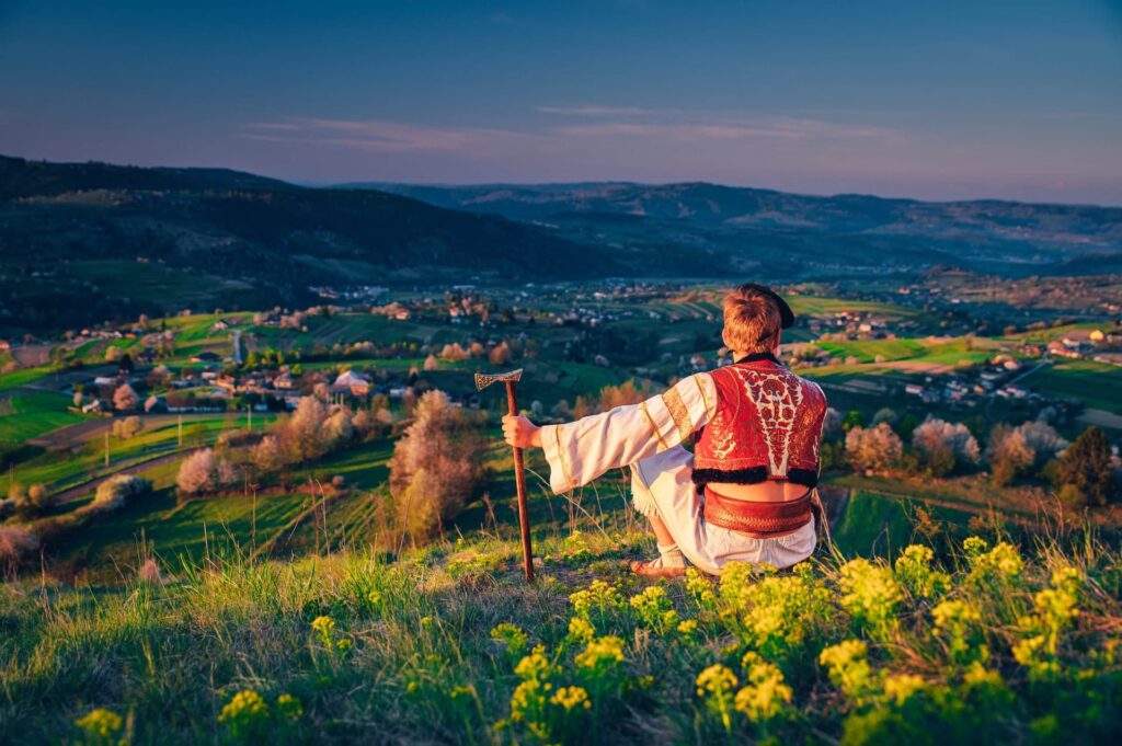 Fotografie zachycuje člověka v tradičním slovenském kroji, který sedí na kopci a dívá se na malebnou krajinu pod ním. Muž drží v ruce pastýřskou hůl a je oblečen v červené vyšívané vestě a bílé košili. Krajina kolem něj je zelená a rozkvetlá, s vesnicí a poli rozprostírajícími se v údolí. V dálce jsou vidět kopce a hory, které se ztrácejí na obzoru. Slunce zapadá, což dodává scéně teplé a zlatavé osvětlení, zvýrazňující krásu slovenské přírody a tradic.