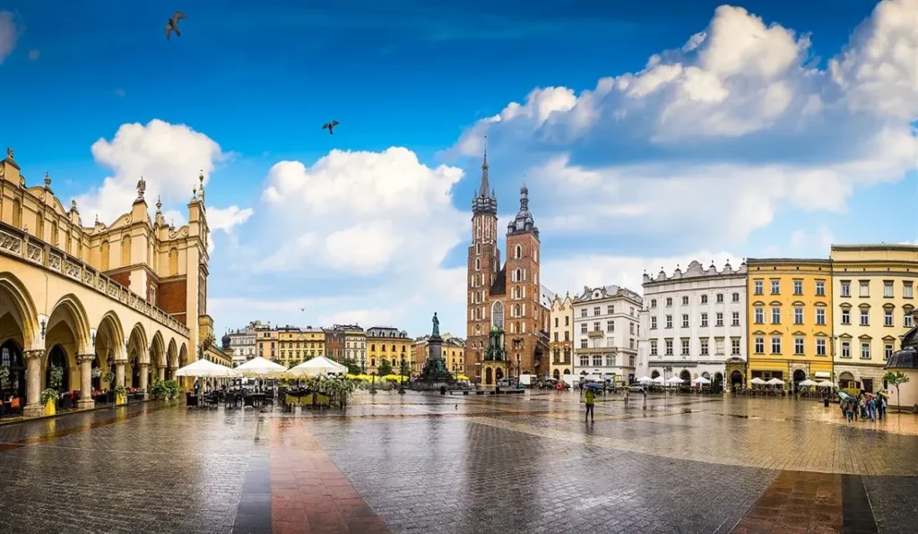 Hlavní náměstí (Rynek Główny) v Krakově, Polsko, po dešti. V popředí jsou deštníky kaváren a restaurací, vlevo historická budova Sukiennice s arkádami, a vpravo Mariánský kostel s dvojitými věžemi. Na obloze jsou bílé mraky a létající ptáci.