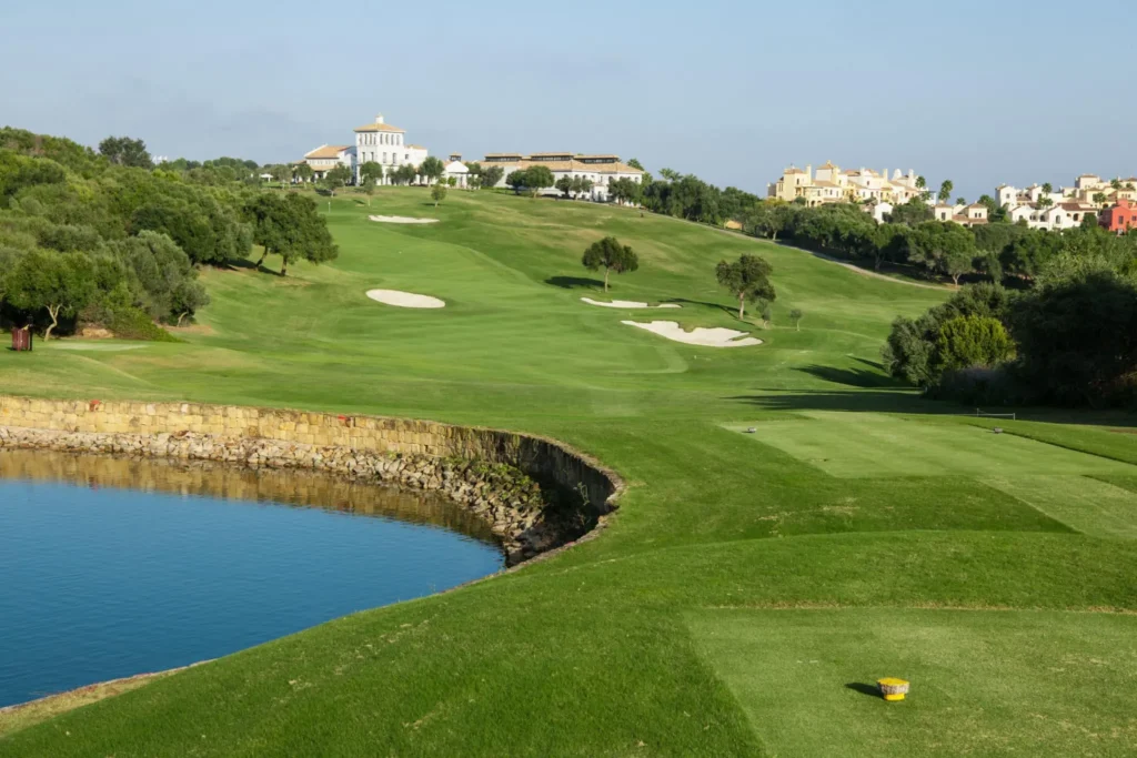 Obrázek zobrazuje La Reserva Golf Club ve Španělsku, jedno z prestižních golfových hřišť v této oblasti. Hřiště je obklopené zelenými fairwayi a pískovými bunkry, které vytvářejí náročné a malebné herní prostředí. V popředí se nachází jezírko s kamenitou hrází, které dodává krajině další prvek krásy. V pozadí jsou vidět elegantní budovy a vilky, které dotvářejí luxusní atmosféru resortu. Celková scenérie je klidná a malebná, s modrou oblohou a příjemným slunečným počasím, ideálním pro golfovou hru.