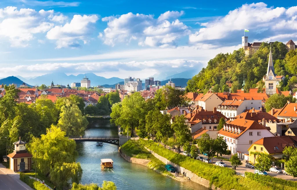 Panorama města Ljubljana, hlavního města Slovinska, s řekou Ljubljanica, starými domy s červenými střechami a Ljubljanským hradem v pozadí na kopci. Na horizontu jsou vidět hory pod modrou oblohou s bílými mraky.