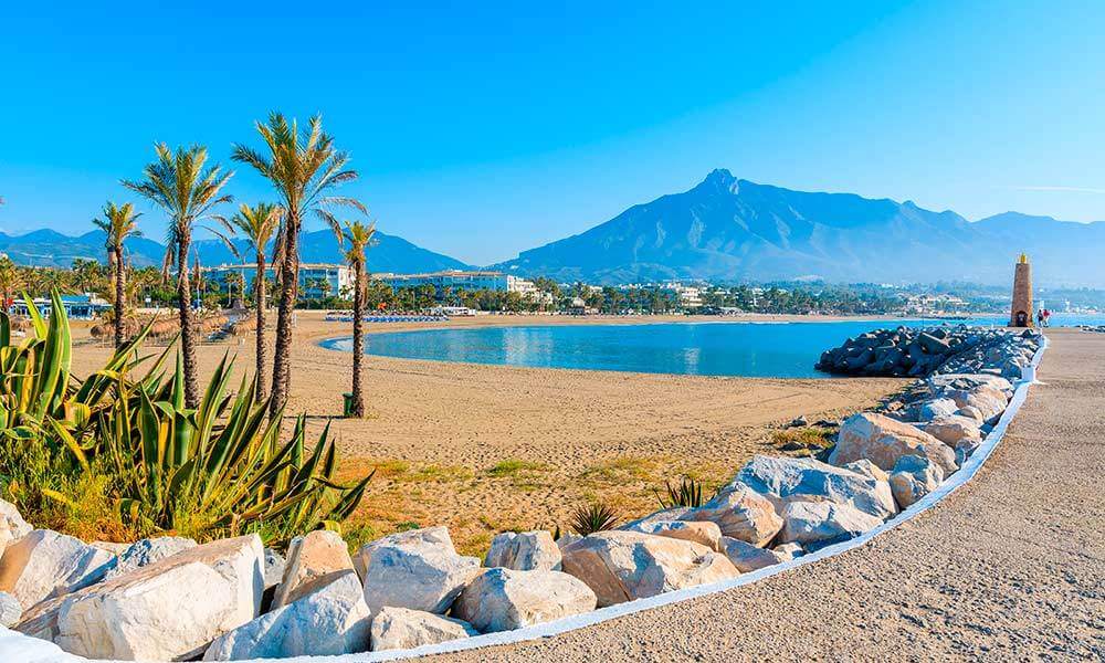 Pohled na nádhernou pláž v Marbelle na Costa del Sol ve Španělsku s modrým mořem, palmovými stromy a horským pozadím. Perfektní místo pro luxusní dovolenou plnou slunce a relaxace.