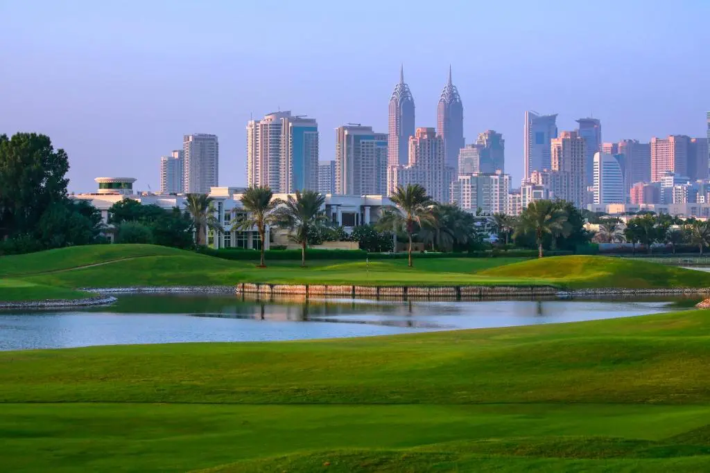 Západ slunce nad Montgomerie Golf Club v Dubaji, který se nachází v těsné blízkosti moderních budov a mrakodrapů. V popředí jsou dobře udržované greeny a vodní překážka, která přidává scenérii na kráse. Palmy a zeleň vytvářejí harmonický kontrast s městským panorama v pozadí, které je zalité jemným večerním světlem, čímž se zvýrazňuje klidná a elegantní atmosféra golfového hřiště.