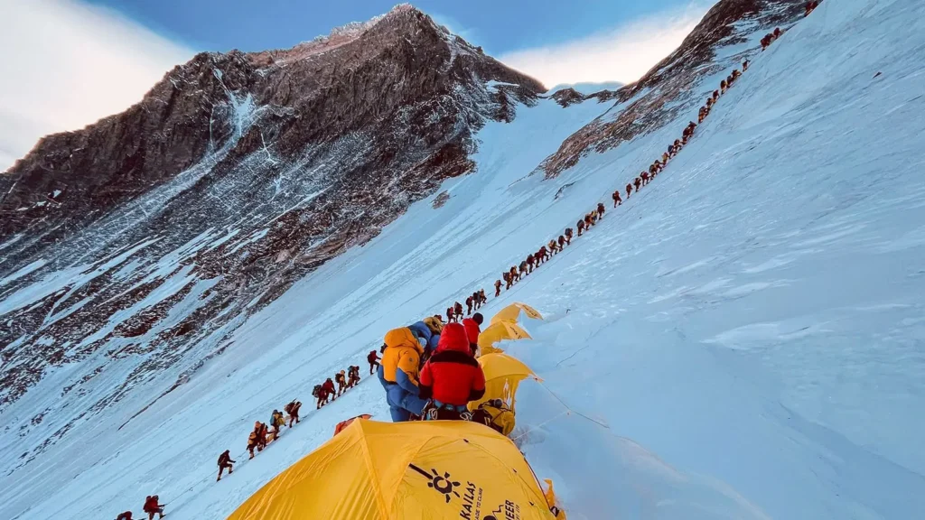 Expedice horolezců v zóně smrti na Mount Everestu, kde horolezci v barevném oblečení šplhají po zasněženém svahu v řadě. Na popředí jsou vidět žluté stany expedičního tábora.