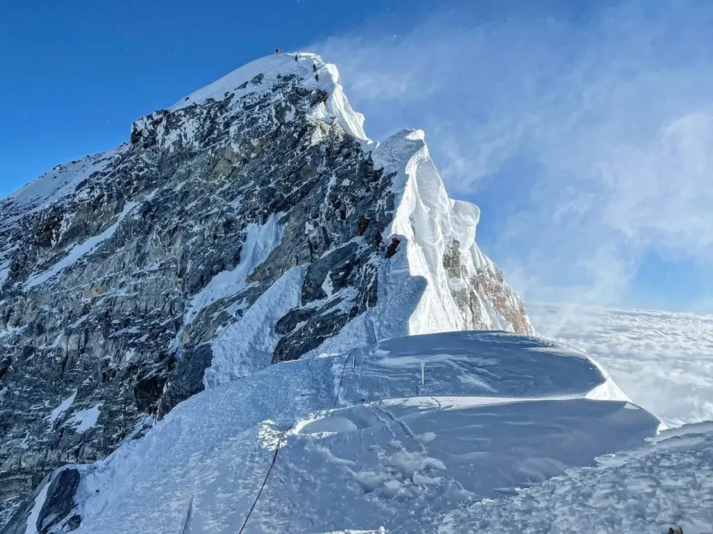 Horolezci na zasněženém hřebenu blízko vrcholu Mount Everestu, nejvyšší hory na světě, stoupají po strmém a ledovém svahu za jasného modrého nebe.