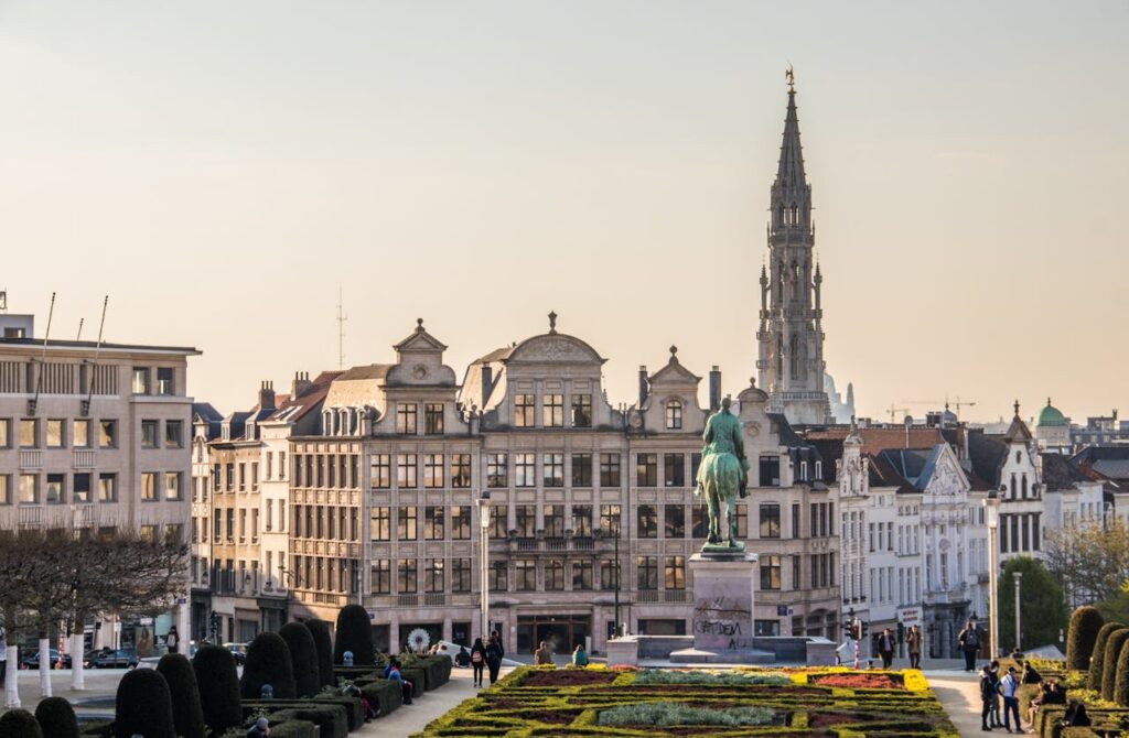 Pohled na historické centrum Bruselu s náměstím Grand Place a gotickou věží radnice v pozadí. V popředí se nachází socha a zahrady, které tvoří krásné místo pro procházky a odpočinek. Okolní budovy ukazují kombinaci historické a moderní architektury.