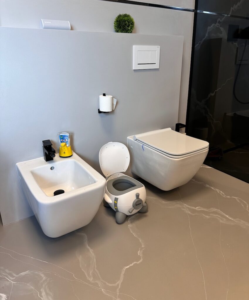 Na obrázku je moderní koupelna se třemi toaletními zařízeními: bidet, záchod a dětský nočník ve tvaru letadla. Na stěně nad záchodem je umístěn toaletní papír a malý zelený keřík jako dekorace. Celý prostor je minimalistický a elegantně zařízený s hladkými povrchy a čistými liniemi.