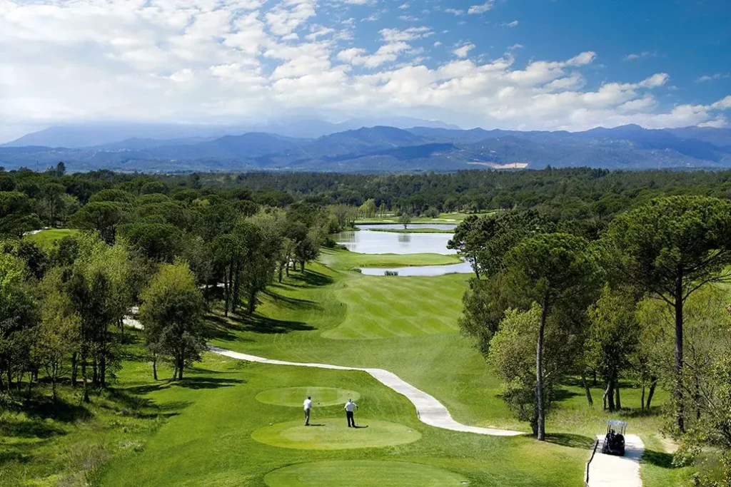 Obrázek zobrazuje PGA Catalunya Resort ve Španělsku, jedno z nejvýznamnějších golfových hřišť v zemi. Hřiště je obklopené hustou zelení a lemované vzrostlými stromy. Dokonale udržované fairwaye vedou hráče kolem několika jezírek, což dodává hřišti přirozenou krásu a náročnost. V pozadí jsou vidět majestátní hory, které přidávají na scenérii. Dva golfisté stojí na odpališti a připravují se na hru. Obloha je modrá s několika obláčky, což vytváří ideální podmínky pro golfový den. Celková atmosféra působí klidně a luxusně.