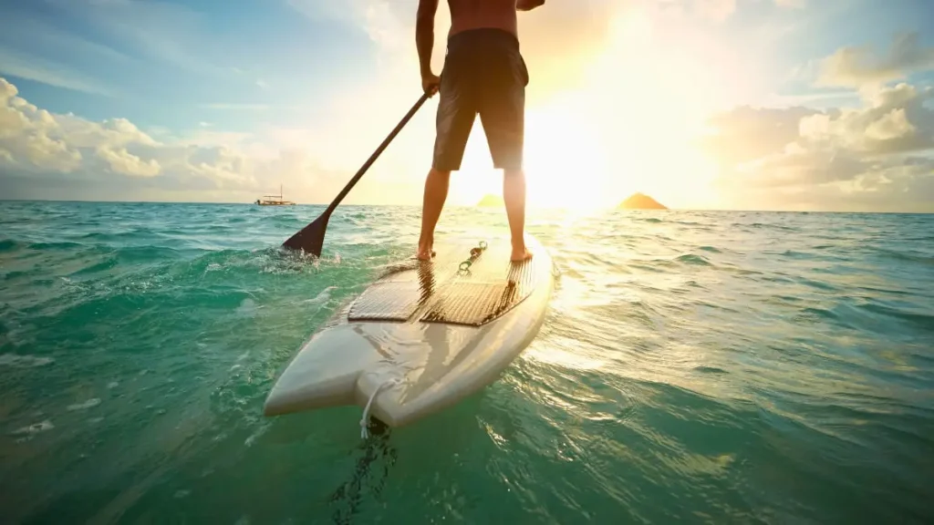 Osoba pádluje na paddleboardu při západu slunce na klidném moři, v pozadí je vidět loď a ostrov.