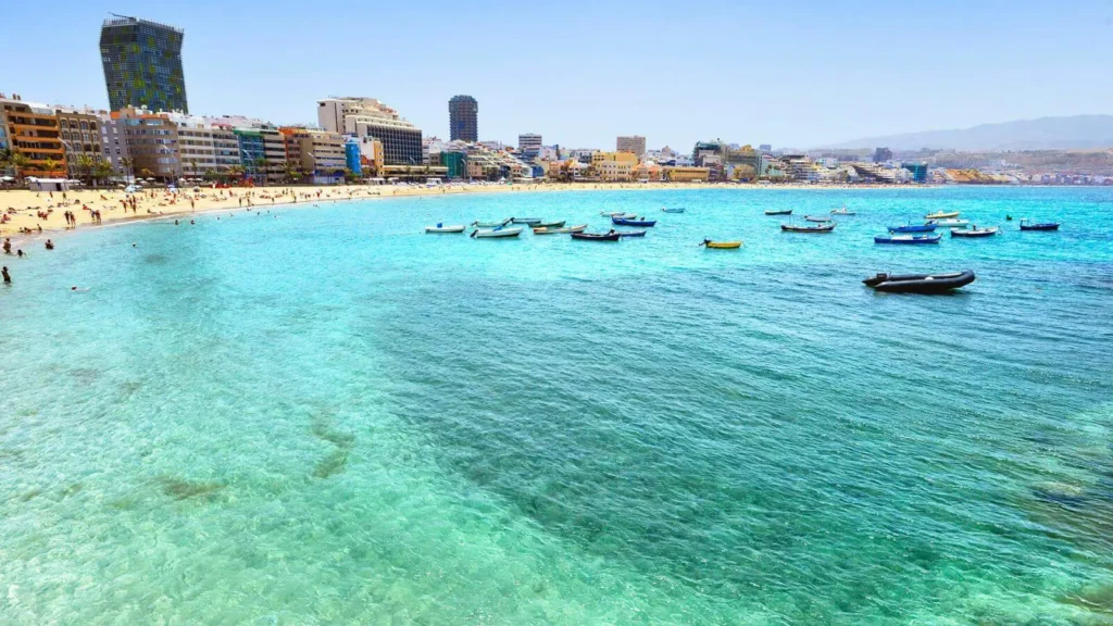 Playa de Las Canteras, jednu z nejznámějších pláží na ostrově Gran Canaria ve Španělsku. Pláž je pokrytá jemným zlatým pískem a obklopená řadou moderních budov a hotelů, které tvoří živou městskou krajinu v pozadí. Moře je čisté a tyrkysové, s několika malými čluny zakotvenými poblíž břehu. Na pláži jsou vidět lidé užívající si slunečného dne, koupající se a relaxující. Obloha je jasná a modrá, což přispívá k celkově klidné a přívětivé atmosféře tohoto populárního letoviska.