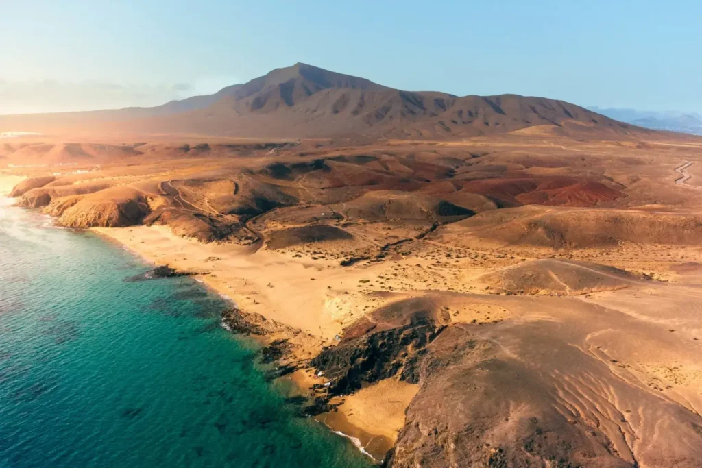 Pohled na pláže na Lanzarote, Kanárské ostrovy, s písčitými plážemi a hornatou sopečnou krajinou v pozadí. Křišťálově čistá voda a zlatavý písek vytvářejí krásný kontrast s červenohnědými kopci.