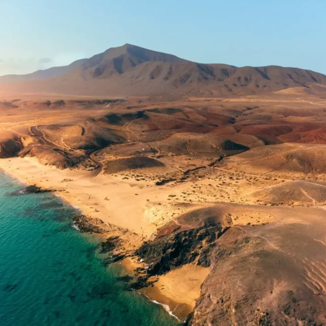 Pohled na pláže na Lanzarote, Kanárské ostrovy, s písčitými plážemi a hornatou sopečnou krajinou v pozadí. Křišťálově čistá voda a zlatavý písek vytvářejí krásný kontrast s červenohnědými kopci.