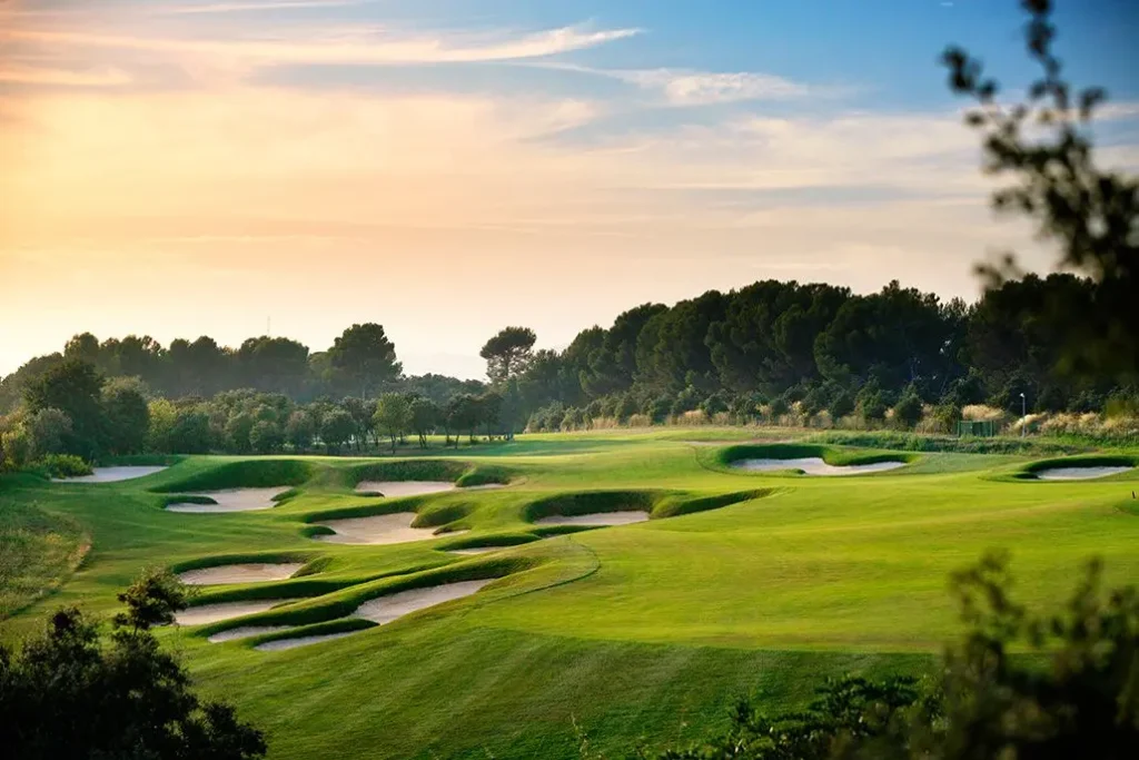 Obrázek zobrazuje Real Club de Golf El Prat v Barceloně, Španělsko. Golfové hřiště je malebně zasazeno do zelené krajiny, s několika dobře udržovanými pískovými bunkry a svěžími fairwayi. Krajina je lemována stromy, které vytvářejí příjemnou a klidnou atmosféru. Na obloze je vidět jemný západ slunce, který dodává scenérii zlatavý nádech a vytváří krásné světelné efekty na trávníku. Celkové prostředí působí poklidně a je ideální pro golfovou hru v nádherné přírodní krajině.