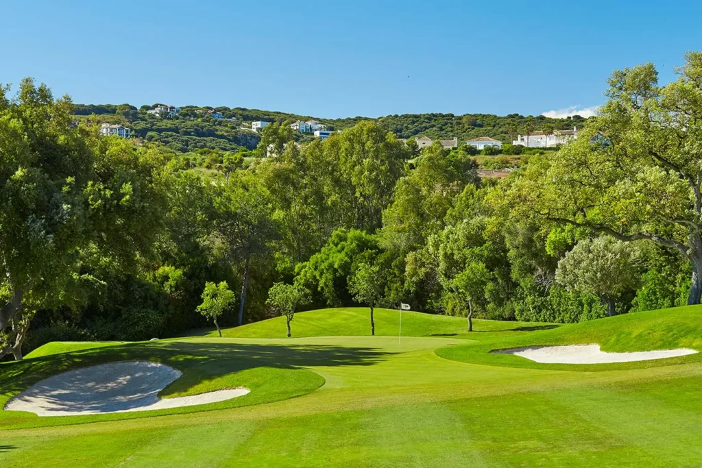 Obrázek zobrazuje část golfového hřiště Real Club de Golf El Prat v Barceloně, jednoho z nejlepších golfových hřišť ve Španělsku. Hřiště je obklopeno bohatou zelení a vzrostlými stromy, které poskytují příjemný stín a estetický zážitek. Pískové bunkry jsou strategicky umístěny kolem greenu, což přidává na výzvě pro hráče. Na vzdáleném horizontu jsou vidět vilky a domy, které se harmonicky začleňují do přírodní krajiny.