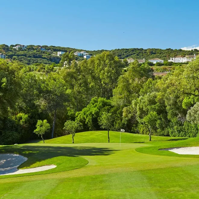 Obrázek zobrazuje část golfového hřiště Real Club de Golf El Prat v Barceloně, jednoho z nejlepších golfových hřišť ve Španělsku. Hřiště je obklopeno bohatou zelení a vzrostlými stromy, které poskytují příjemný stín a estetický zážitek. Pískové bunkry jsou strategicky umístěny kolem greenu, což přidává na výzvě pro hráče. Na vzdáleném horizontu jsou vidět vilky a domy, které se harmonicky začleňují do přírodní krajiny.