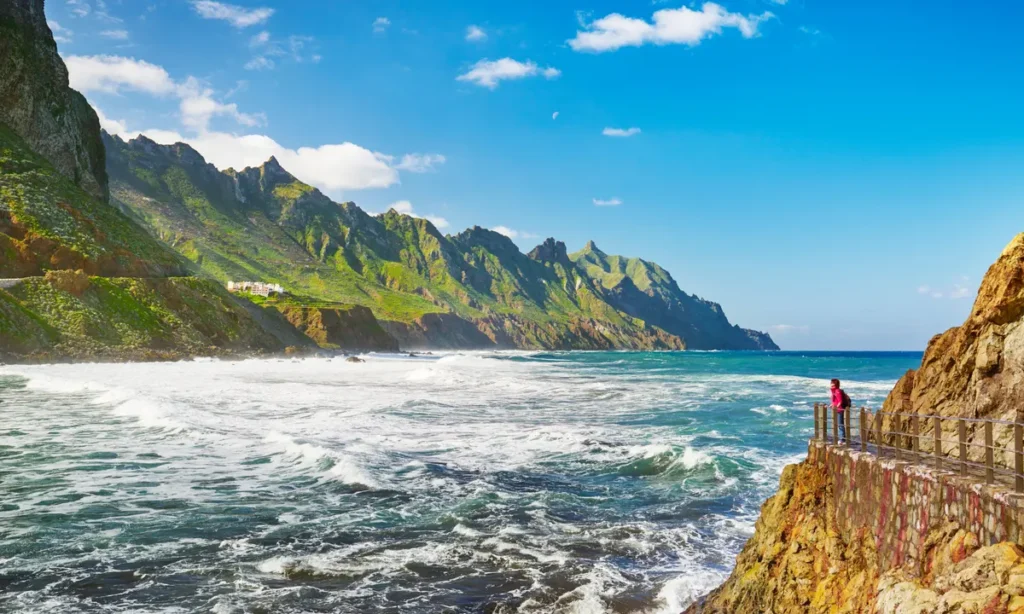 Pohled na dramatické pobřeží Tenerife s divokými vlnami, strmými zelenými horami a turistou stojícím na vyhlídce nad skalnatým útesem.