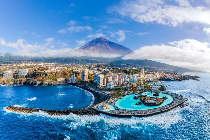 Letecký pohled na pobřeží Tenerife s městem, bazénovým komplexem a majestátním vrcholem sopky Teide v pozadí, částečně zahaleným mraky.