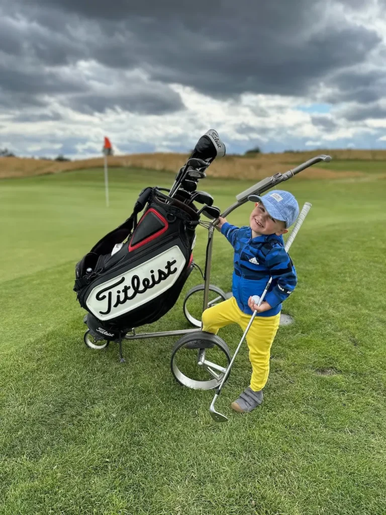 Malý chlapec stojí vedle golfového vozíku s golfovou taškou značky Titleist na golfovém hřišti. Chlapec má na sobě modrou bundu, žluté kalhoty a čepici, zatímco se usmívá na kameru. V pozadí je golfový green s vlajkou a dramatická obloha s tmavými mraky.