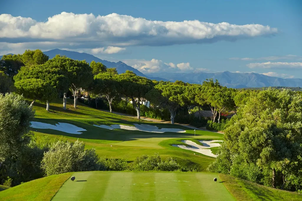 Obrázek zobrazuje Valderrama Golf Club ve Španělsku. Golfové hřiště je obklopené hustou zelení a stromy, s dokonale udržovanými fairwaye a pískovými bunkry. V pozadí jsou vidět hory, které dodávají krajině majestátní vzhled. Obloha je jasně modrá s několika bílými oblaky, což naznačuje krásný slunečný den ideální pro golf. Celkové prostředí působí klidně a malebně, což je typické pro jedno z nejprestižnějších golfových hřišť ve Španělsku.