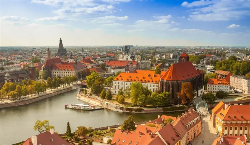 Panoramatický pohled na historické centrum Wroclawi, Polsko. Viditelné jsou červené střechy starých budov, řeka Odra s lodí a zelené parky. V pozadí se tyčí gotická architektura a moderní budovy, které dotvářejí městskou scenérii pod modrou oblohou.