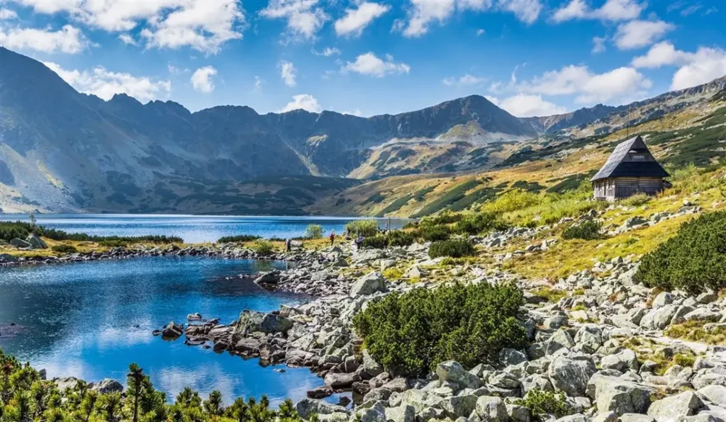 Vysoké Tatry v Polsku, s krásným horským jezerem a skalnatým pobřežím. V pozadí se tyčí majestátní hory, zatímco v popředí se nachází tradiční dřevěná horská chatka. Slunečný den s modrou oblohou a obláčky dodává scenérii poklidnou atmosféru.