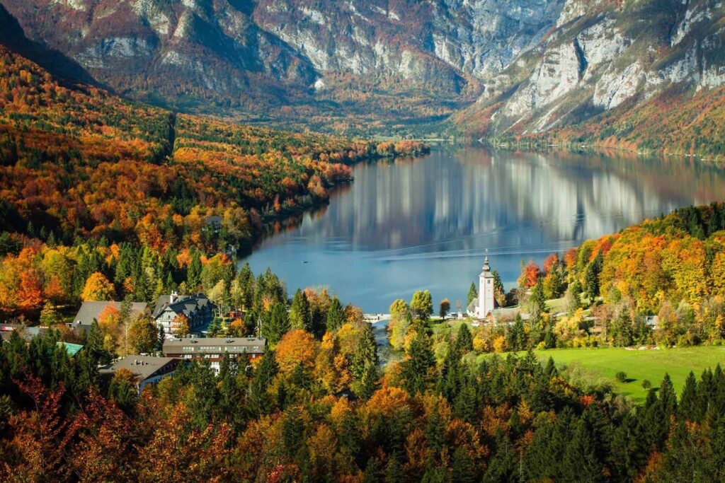 Jezero Bohinj v Triglavském národním parku, Slovinsko, obklopené hustými lesy v podzimních barvách a vysokými horami v pozadí. V popředí je malá vesnice s kostelem a okolní zelené louky.