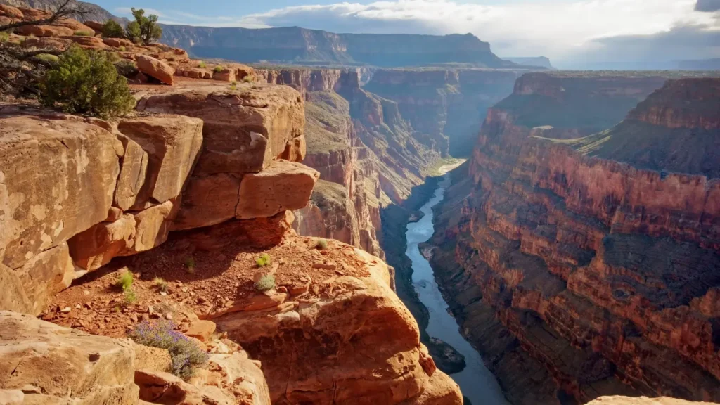 Pohled na hluboké údolí Grand Canyonu s řekou Colorado, která se vine mezi strmými červenými skalami. V popředí jsou robustní skalní útvary a zelené keře, které dodávají kontrast této dramatické krajině.