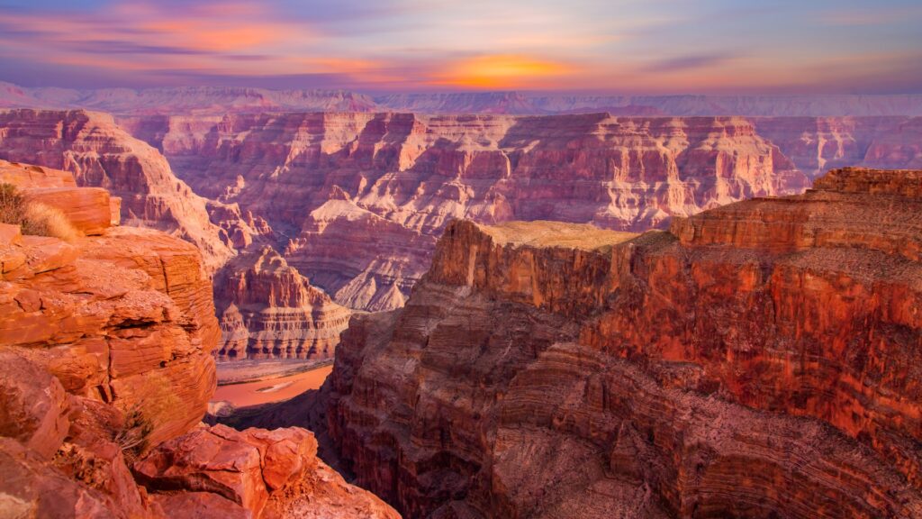 Pohled na Grand Canyon při západu slunce, kdy červené a oranžové vrstvy hornin září v měkkém světle a odhalují nádherné detaily geologických útvarů a hlubokých údolí.