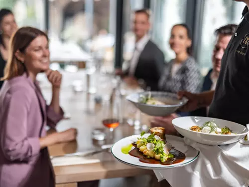 Obsluha servíruje jídlo skupině lidí sedících u stolu v elegantní restauraci.