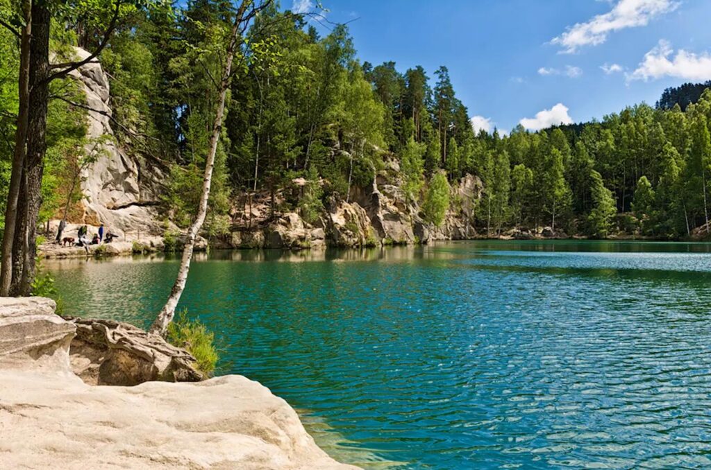 Křišťálově čisté jezero obklopené skalními útvary a hustými lesy v oblasti Adršpašsko-teplických skal. V popředí je jemně nakloněný strom, zatímco na pozadí se rýsují impozantní pískovcové skály. Klidná vodní hladina odráží modrou oblohu a okolní zeleň, vytvářejíce tak idylickou přírodní scenérii.