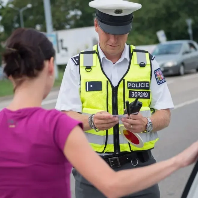 Policista v reflexní vestě provádí silniční kontrolu a kontroluje doklady ženy řidičky, která stojí u svého auta.