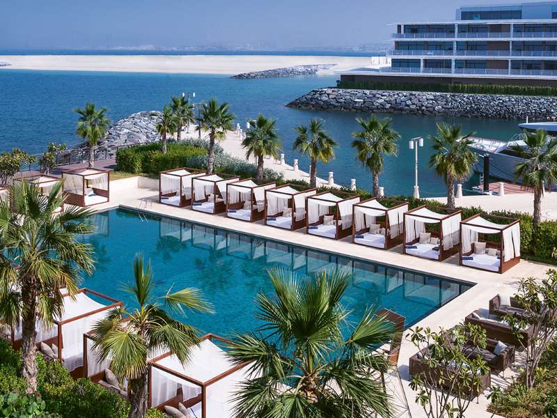 Pohled na elegantní bazén obklopený pohodlnými lehátky a palmami v Bulgari Resort Dubai, s výhledem na oceán.