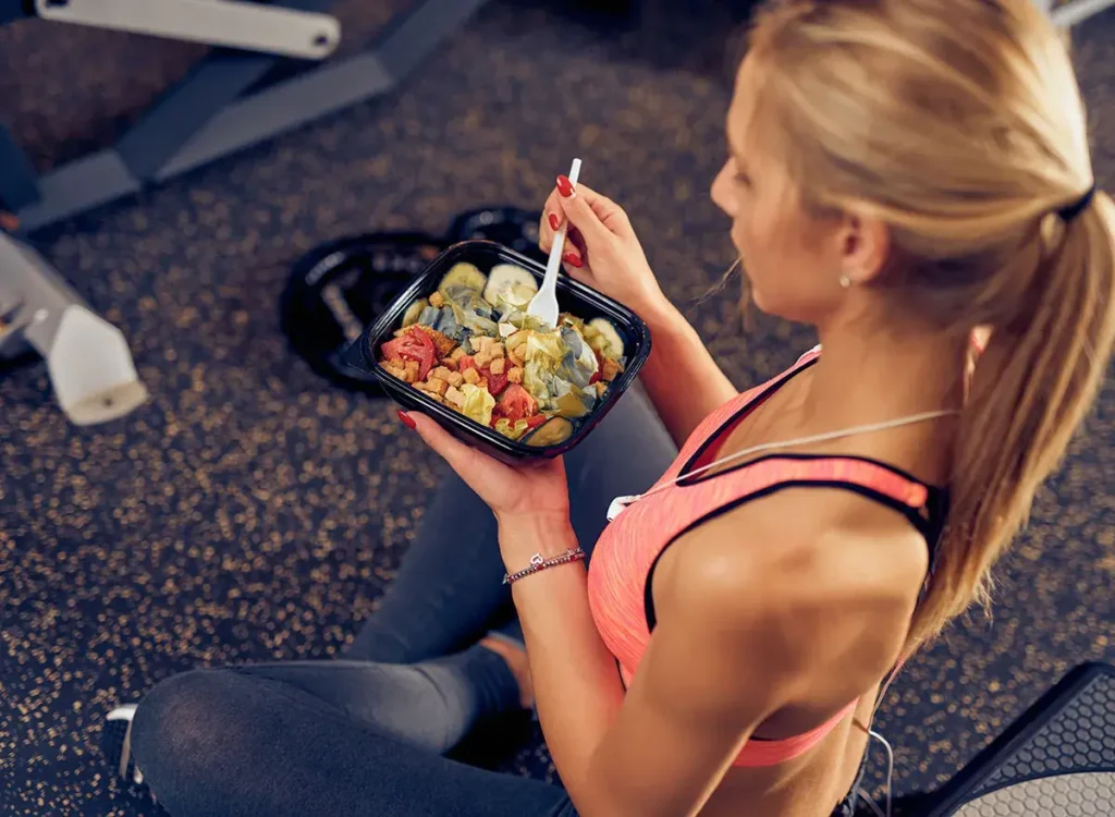 Žena sedící ve fitness centru, držící misku se zdravým jídlem před tréninkem. Má na sobě sportovní oblečení a lžíci, kterou jí pokrm bohatý na zeleninu a bílkoviny.