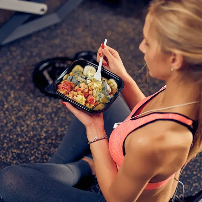 Žena sedící ve fitness centru, držící misku se zdravým jídlem před tréninkem. Má na sobě sportovní oblečení a lžíci, kterou jí pokrm bohatý na zeleninu a bílkoviny.