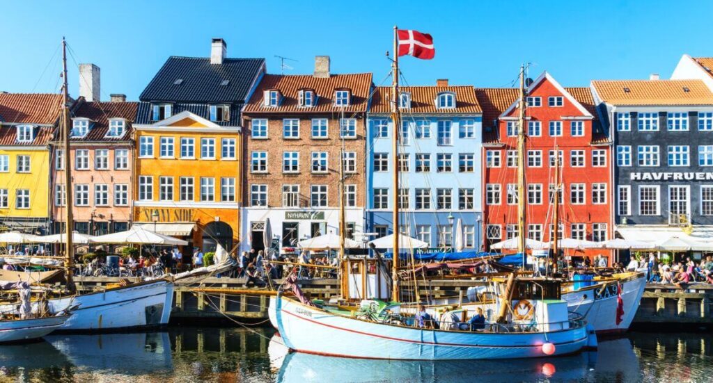 Malebný přístav Nyhavn v Kodani, Dánsko, s barevnými historickými domy, loděmi na klidné vodní hladině a dánskou vlajkou vlající na stožáru. Nyhavn je jednou z nejznámějších a nejnavštěvovanějších turistických atrakcí v dánském hlavním městě.