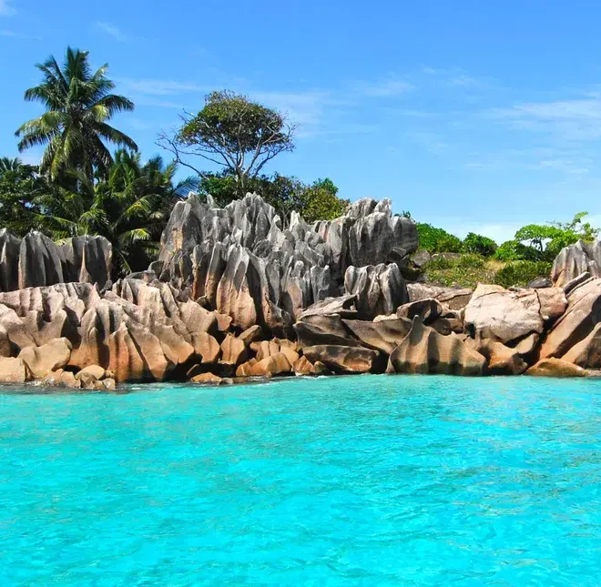 Pohled na tyrkysovou vodu a žulové skaliska na Seychelách, obklopené palmami a tropickou vegetací.