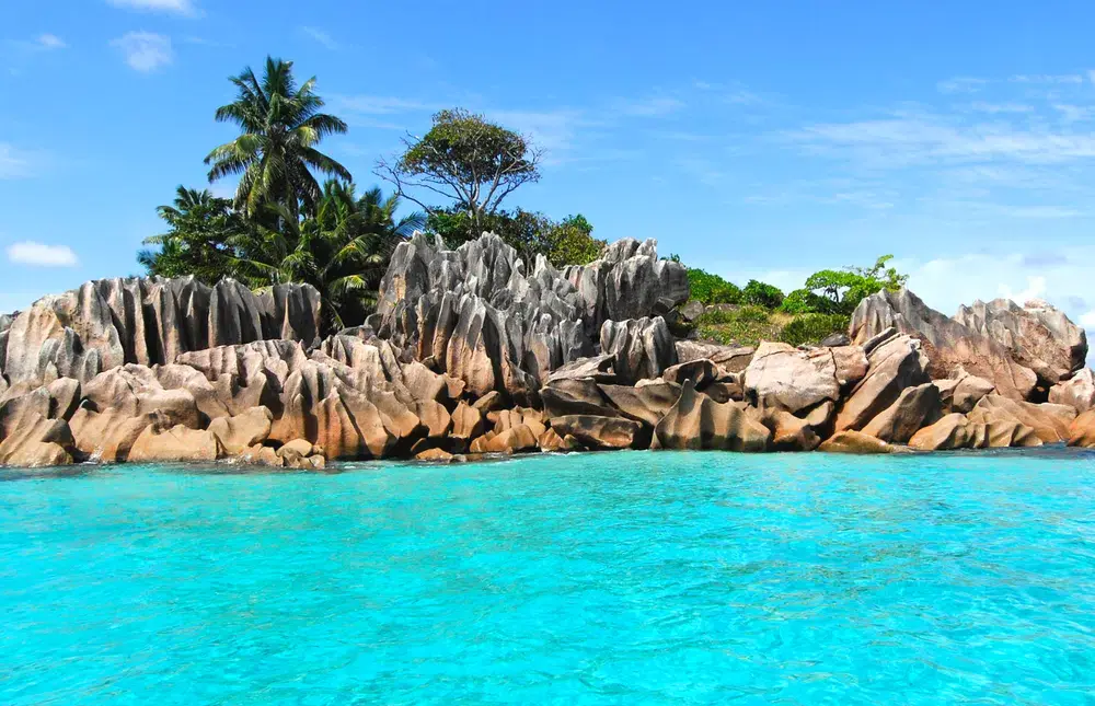 Pohled na tyrkysovou vodu a žulové skaliska na Seychelách, obklopené palmami a tropickou vegetací.