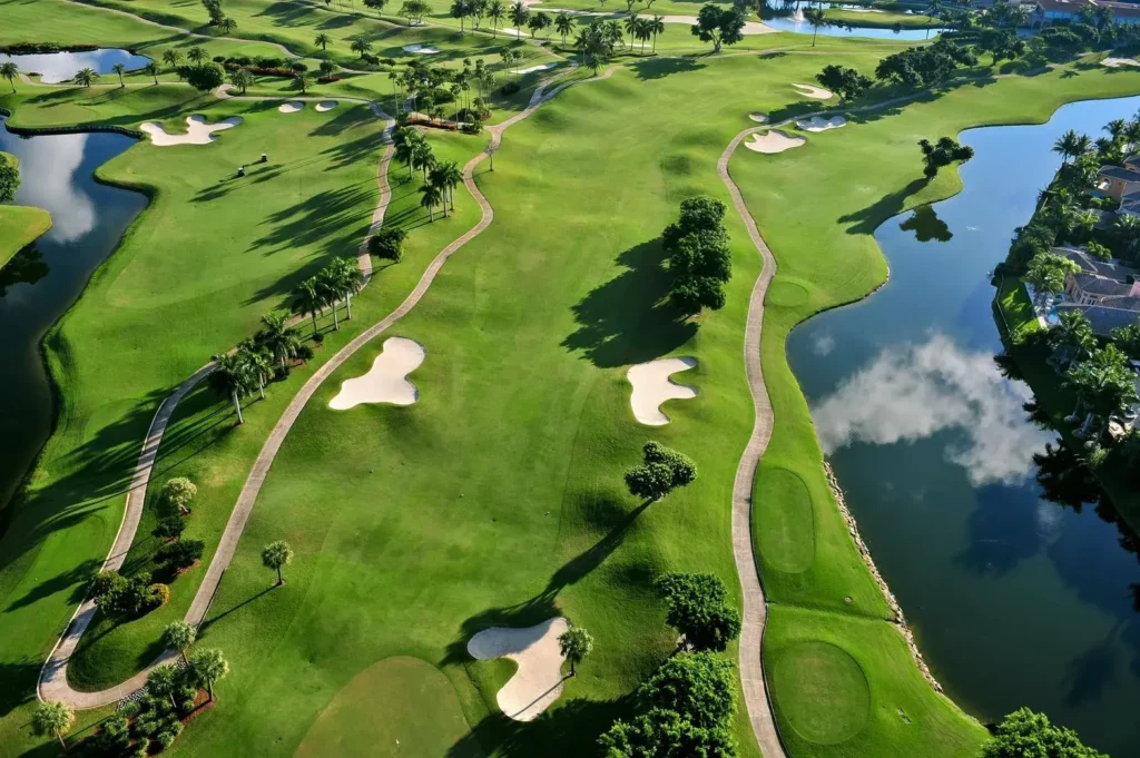 Letecký pohled na rozlehlé golfové hřiště obklopené zelení, s několika jezery a vodními překážkami. Na hřišti jsou vidět písečné bunkry, stromy a klikaté cesty vedoucí mezi jednotlivými jamkami. Vpravo jsou patrné budovy u vody, které tvoří malebné pozadí tohoto sportovního areálu.