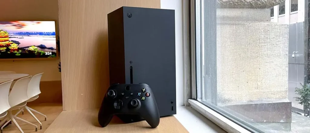 Herní konzole Microsoft Xbox Series X s ovladačem na dřevěné polici vedle okna.